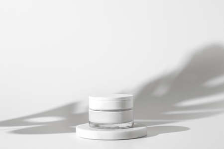 White unbranded cosmetic cream jar standing on white podium. Skin care product presentation on the white background with shadow of exotic plant. Trendy mockup. Skincare, beauty and spa.の写真素材