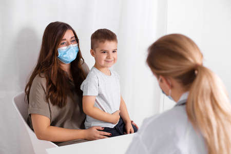 Young brunette woman with little boy having consultation at pediatrician office. Doctor, child and mother wearing facemasks during flu outbreak. Virus protection.の写真素材
