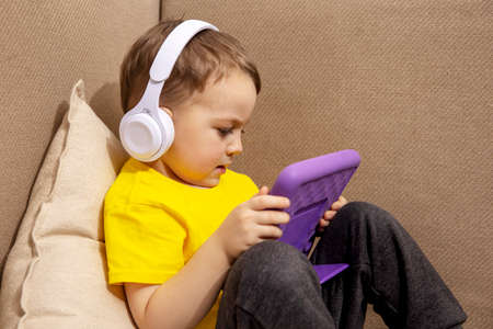 Little caucasian boy with yellow shirt playing game on digital tablet at home. Portrait of a child at home watching cartoon on violet tablet. Modern kid and education technology.の写真素材