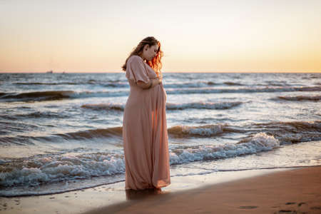 Young pregnant woman with a beautiful sea view on the background. Happy and calm pregnant woman with long hair and pink dress standig on the beach. Romantic view, ocean, sunset, maternity.の写真素材