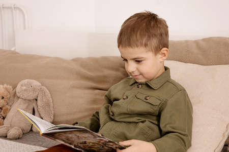 Little and cute caucasian boy reading a book on the bed at home. Interior and clothes in natural...