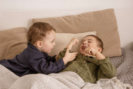 Two little and cute caucasian boys playing together on the bed at home. Interior and clothes in natural earth colors. Cozy environment. Children having fun, two brothers kittle each other.の写真素材