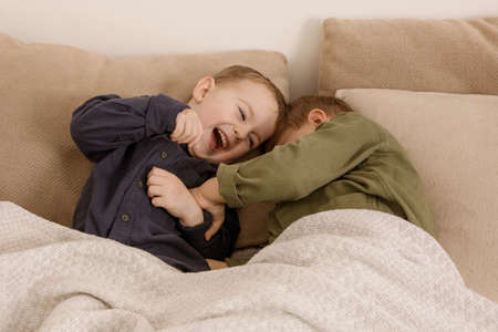 Two little and cute caucasian boys playing together on the bed at home. Interior and clothes in natural earth colors. Cozy environment. Children having fun, two brothers kittle each other.の写真素材