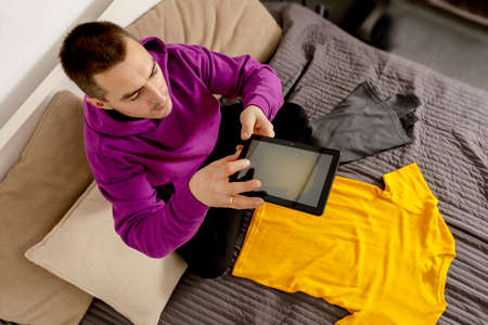 Man with violet hoodie using digital tablet, takes a photo of his old clothes to sell them online....