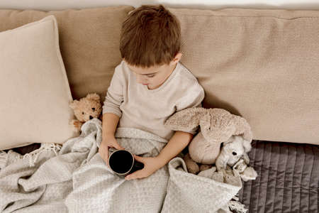 Sick little boy drinking hot tea on the bed at home. Unwell, illness child wrapped in a blanket, with mug in his room. Flu season. Interior and clothes in natural earth colors. Cozy environment.の写真素材