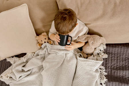 Sick little boy drinking hot tea on the bed at home. Unwell, illness child wrapped in a blanket, with mug in his room. Flu season. Interior and clothes in natural earth colors. Cozy environment.の写真素材