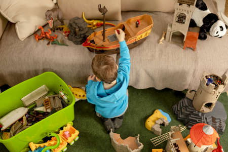 Little caucasian boy with blue hoodie playing with colourful toys at home. Child having fun. Happy and cheerful kid plays with ship, dinosaurs, castle. Leisure activity, domestic life. Cozy room.の写真素材