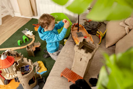 Little caucasian boy with blue hoodie playing with colourful toys at home. Child having fun. Happy and cheerful kid plays with ship, dinosaurs, castle. Leisure activity, domestic life. Cozy room.の写真素材