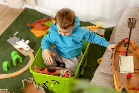 Little caucasian boy with blue hoodie playing with colourful toys at home. Child having fun. Happy and cheerful kid plays with ship, dinosaurs, castle. Leisure activity, domestic life. Cozy room.の写真素材