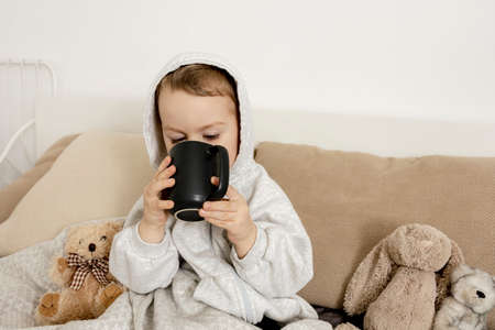 Sick little boy drinking hot tea on the bed at home. Unwell, illness child wrapped in a blanket, with mug in his room. Flu season. Interior and clothes in natural earth colors. Cozy environment.の写真素材