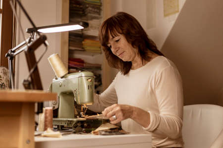Caucasian brunette woman sewing fabric with a vintage, retro sewing machine. Fashion, creation and tailoring. Process of sewing in atelier or workshop. Special hobby. Cozy interior and warm colors.の写真素材