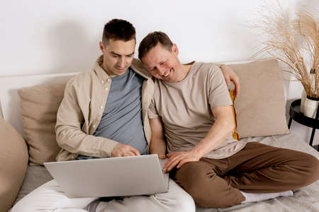 Happy gay couple with casual clothes spending time together at home and watching movie on the laptop. Two caucasian men relaxing. Homosexual relationships and alternative love. Cosy interior.の写真素材