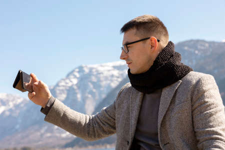 Young adult man sitting outdoors on bench and making selfie with his smartphone, enjoying mountains,...