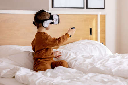 Little adorable boy sitting on bed at home with VR headset and playing interactive video game,...