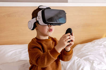 Little adorable boy sitting on bed at home with VR headset and playing interactive video game,...