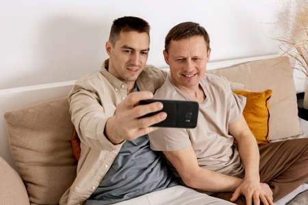 Happy gay couple with casual clothes spending time together at home and making selfie on smartphone....
