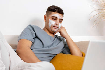 Young man with casual clothes and beauty mask on the face lying on bed with laptop computer and resting. Cosmetic for men, skin care. Man reading news in internet or watching movie. Relaxing at home.の写真素材
