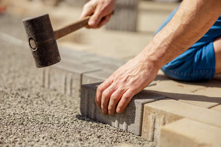 Young man laying gray concrete paving slabs in house courtyard on gravel foundation base. Master lays paving stones. Garden brick pathway paving by professional paver worker. Repairing sidewalk.の写真素材