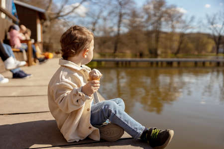 Little adorable boy sitting outdoors and eating ice cream. Lake, water and sunny weather. Child and sweets, sugar. Kid enjoy a delicious dessert. Preschool child with casual clothing. Positive emotionの写真素材