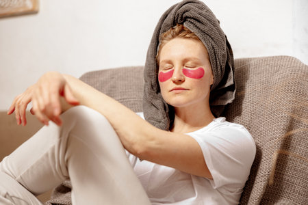 Young woman with casual clothes, towel on head and eye patches sitting on couch and resting. Relax after taking a shower. Cosmetic for women, skin care. Self care, time for yourself. Relaxing at homeの写真素材