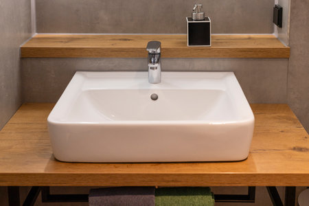 A white ceramic sink sits on a wooden countertop in a contemporary bathroom. The faucet is polished chrome, reflecting the ambient light in a streamlined settingの写真素材