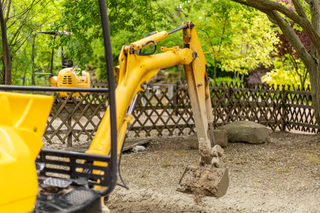 A yellow excavator is digging into the soil at a construction site surrounded by green trees and a wooden fence, highlighting a busy outdoor work environmentの写真素材