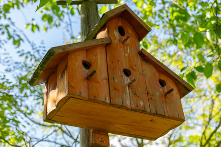 A beautifully crafted wooden birdhouse with multiple openings is suspended from a wooden pole....