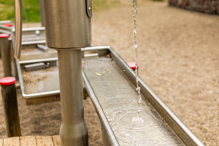 A metal water pump is actively dispensing clear water into a long channel designed for children. The...