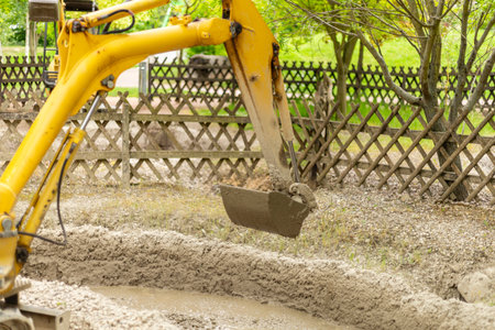 A yellow excavator performs earthmoving tasks in a construction zone surrounded by a wooden fence. The machinery lifts a bucket filled with soil under clear skiesの写真素材