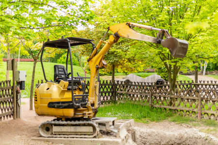 A yellow excavator is parked at a construction site with a wooden fence nearby. Lush green trees provide a backdrop under clear skies, indicating daytime activities at the locationの写真素材
