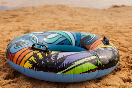 An inflatable tube with vibrant patterns sits on a sandy beach, ready for use on a warm day. The...