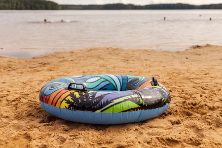 A vibrant inflatable tube sits on golden sand beside a lake. Swimmers can be seen enjoying the clear...