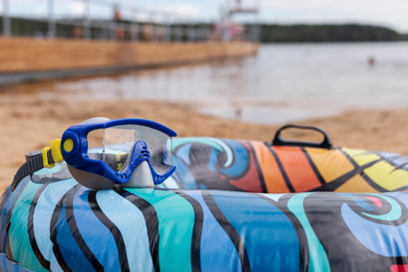 Brightly colored inflatable ring rests on the sandy shore of a lake, paired with snorkeling goggles. The cloudy sky looms over calm waters, creating a serene atmosphere for relaxationの写真素材