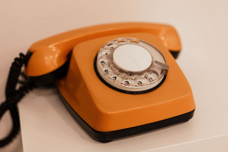 A bright orange rotary telephone is positioned on a white table, highlighting its retro design. This...