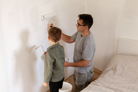 A father and his young son work together to paint the walls of their bedroom. They are using rollers to apply fresh white paint, creating a bright atmosphereの写真素材