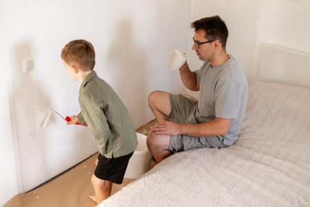 A man sits on the bed sipping coffee as his young son helps paint the bedroom walls. They are both...