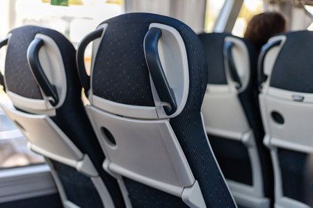 Passengers travel inside a bus, viewing the backs of comfortable seats with handles. Sunlight...