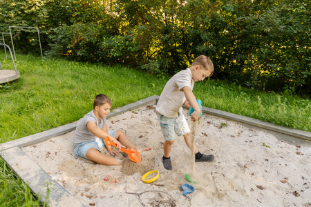 Two children are happily playing in a sandbox, digging with colorful shovels. Lush green grass and trees create a natural backdrop in a warm afternoon setting, enhancing their funの写真素材