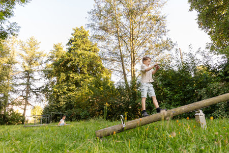 Two children engage in playful activities in a lush green park during daylight. One child balances on a log, while the other enjoys the surrounding natureの写真素材
