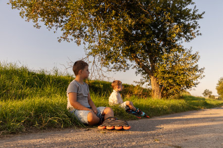 Two children sit on a grassy area, taking a break from inline skating. They are enjoying their time outdoors on a sunny afternoon, surrounded by trees and natureの写真素材