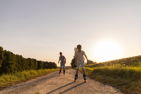 Two friends are having fun inline skating down a dirt path surrounded by greenery under a clear blue sky during late afternoon sunshineの写真素材