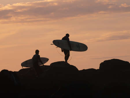 Two surfers walk with surfboards over the rocks to the sea during sunsetの写真素材
