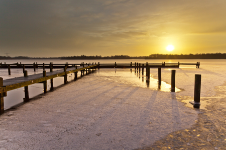 Amber Colored Sunrise above a Jetty on a Frozen Winter Lake with Ice and Snowの写真素材