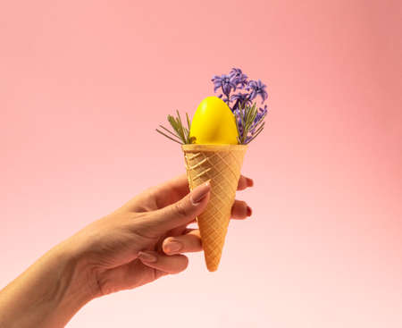 Hand holding ice cream cone with yellow egg and spring flowers hyacinths on pink background. Retro...