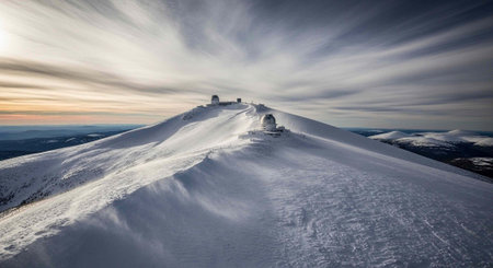 A snow-covered mountain peak with buildings at the summit under a cloudy sky during sunset or...