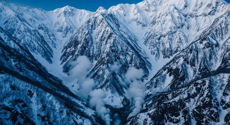 Aerial view of rugged snow-covered mountains with clouds and a clear blue sky