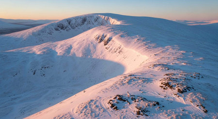 Aerial view of snow-covered mountains with gentle slopes and clear blue sky at dawn or dusk.