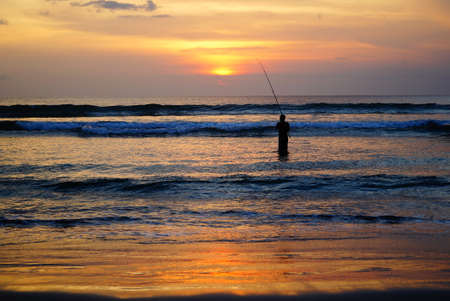 Silhouette of fisherman standing with fishing rod in the sea at sunset. Bali. Indonesia.の写真素材