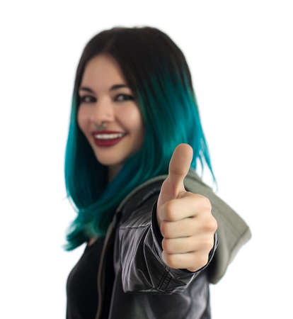 Smiling girl showing the thumb up sign on a white background. Pierced, turquoise haired and dressing a leather jacket.の写真素材