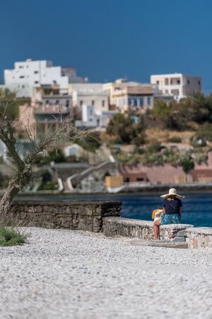 Syros island, Cyclades archipelago, Greece - A woman with white hat is sitting on a wall and looking at the seaの写真素材